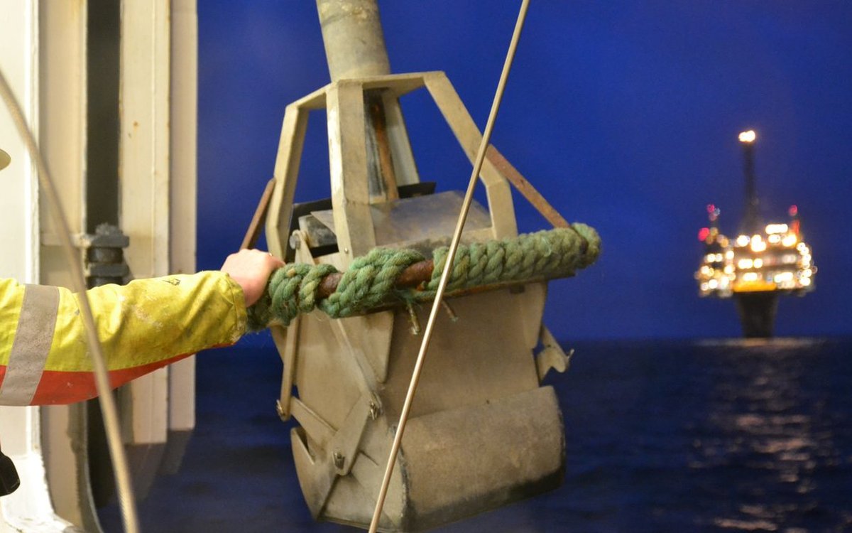 En grabb som blir brukt til å hente opp prøver av havbunnen blir tatt om bord på en båt. I bakgrunnen er det en oljeplattform.