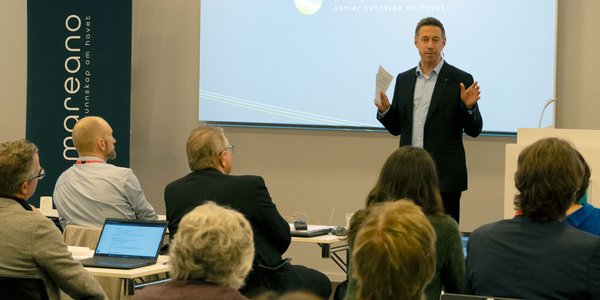 En mann i dress står foran publikum, han er engasjert og snakker til de.